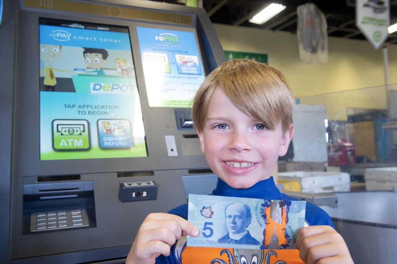 A smiling boy at a bottle depot holding up a five-dollar bill